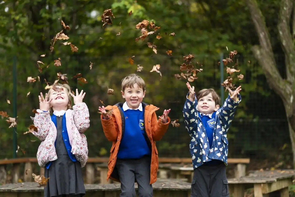 Three children in Stedham fleeces and coats throwing armfuls of autumn leaves into the air
