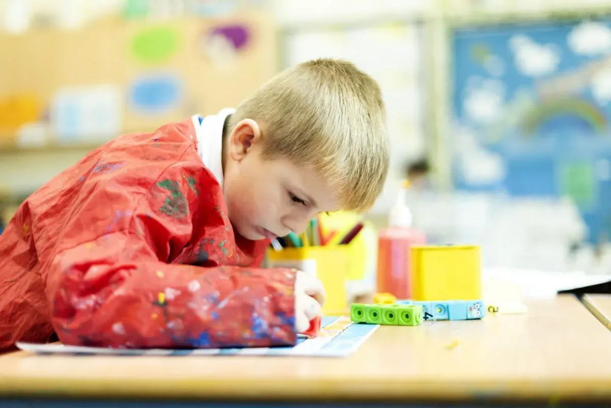 A young pupil in a red apron concentrating on drawing at a table