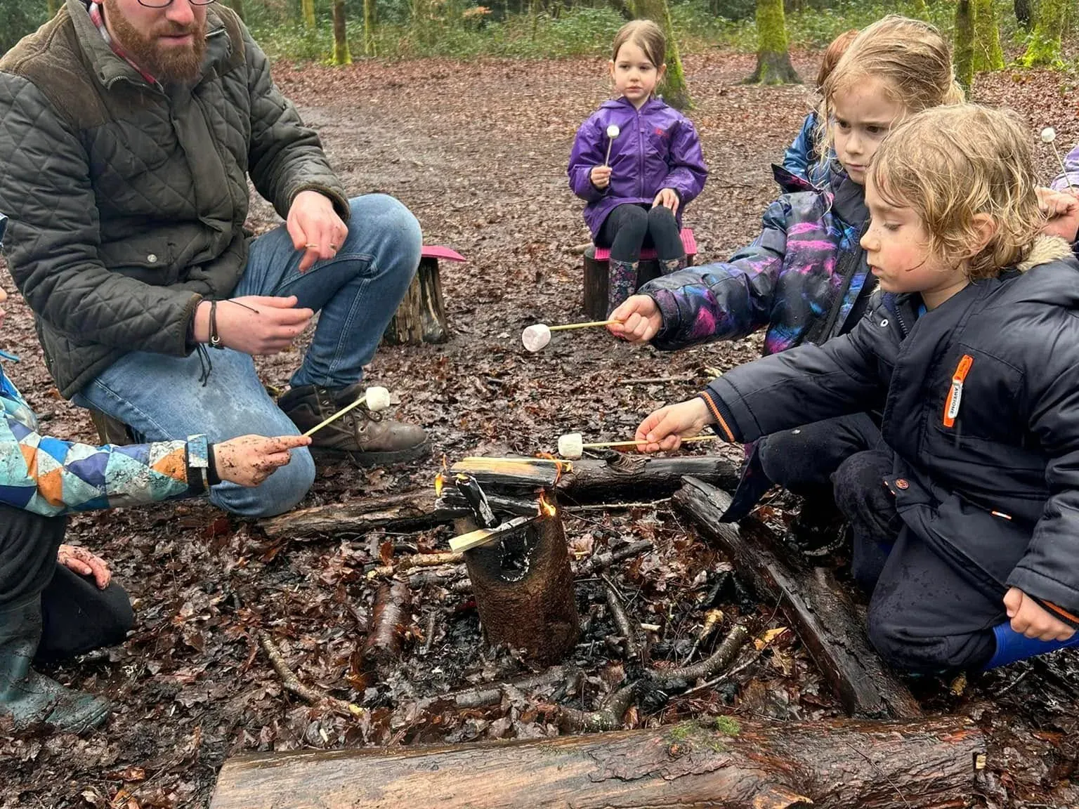 A teacher and children sitting on logs around a campfire in the woods, toasting marshmallows on sticks