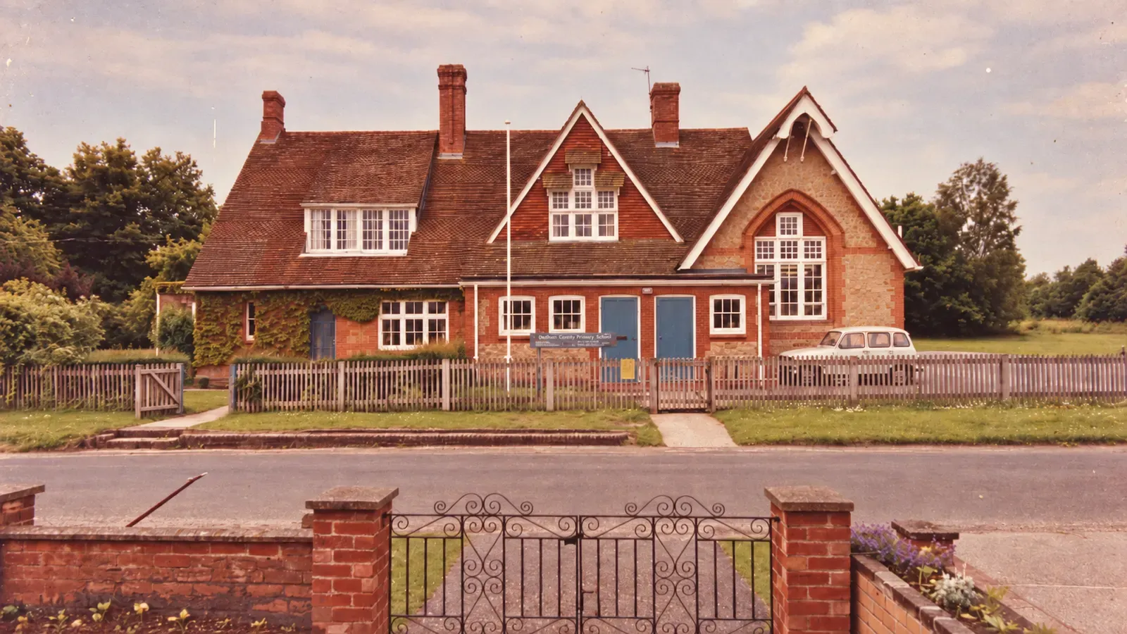 Stedham County Primary School from the road, with a wooden picket fence in front and wrought-iron gates in the foreground