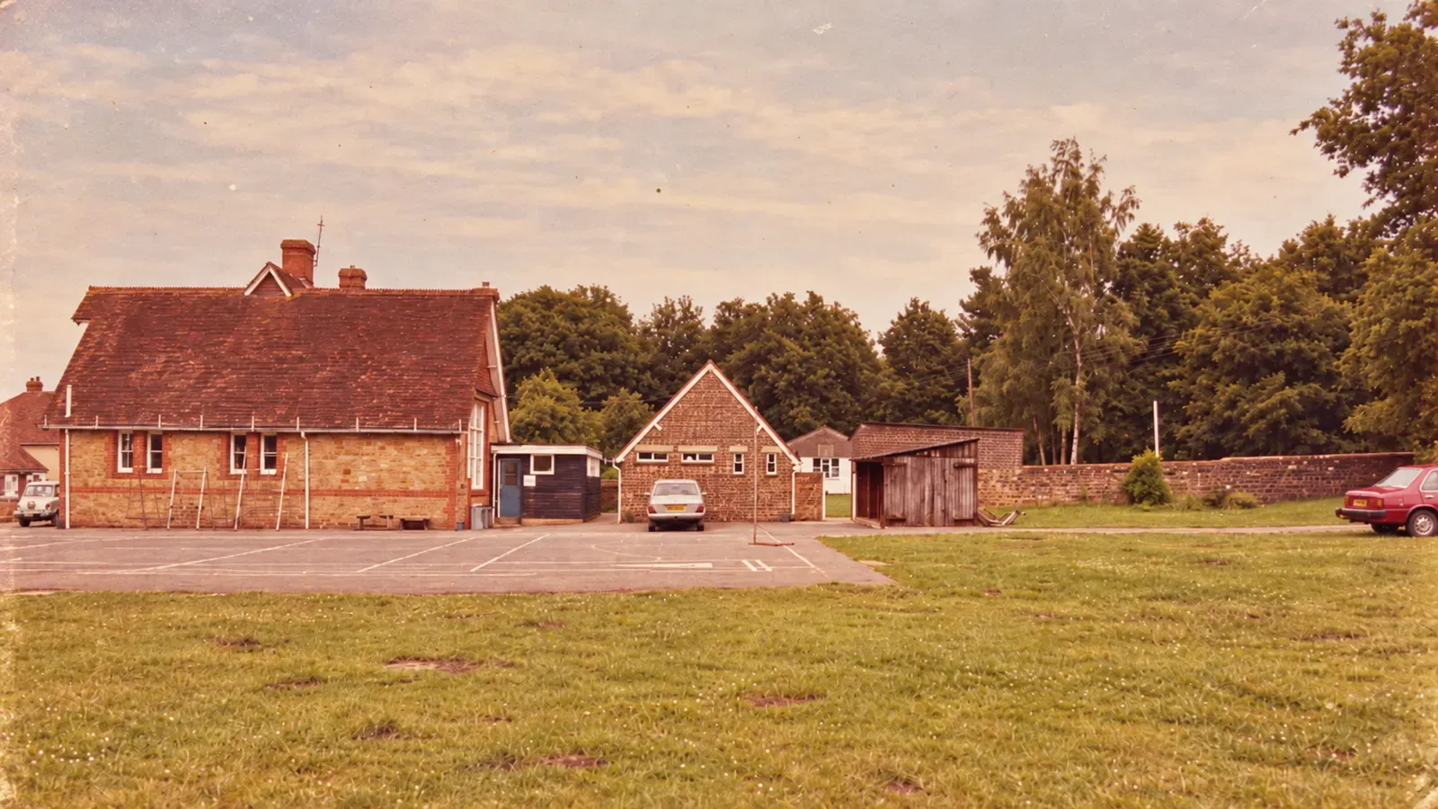 The playground and the back of the school buildings, with a car parked on the tarmac and a grass field in front