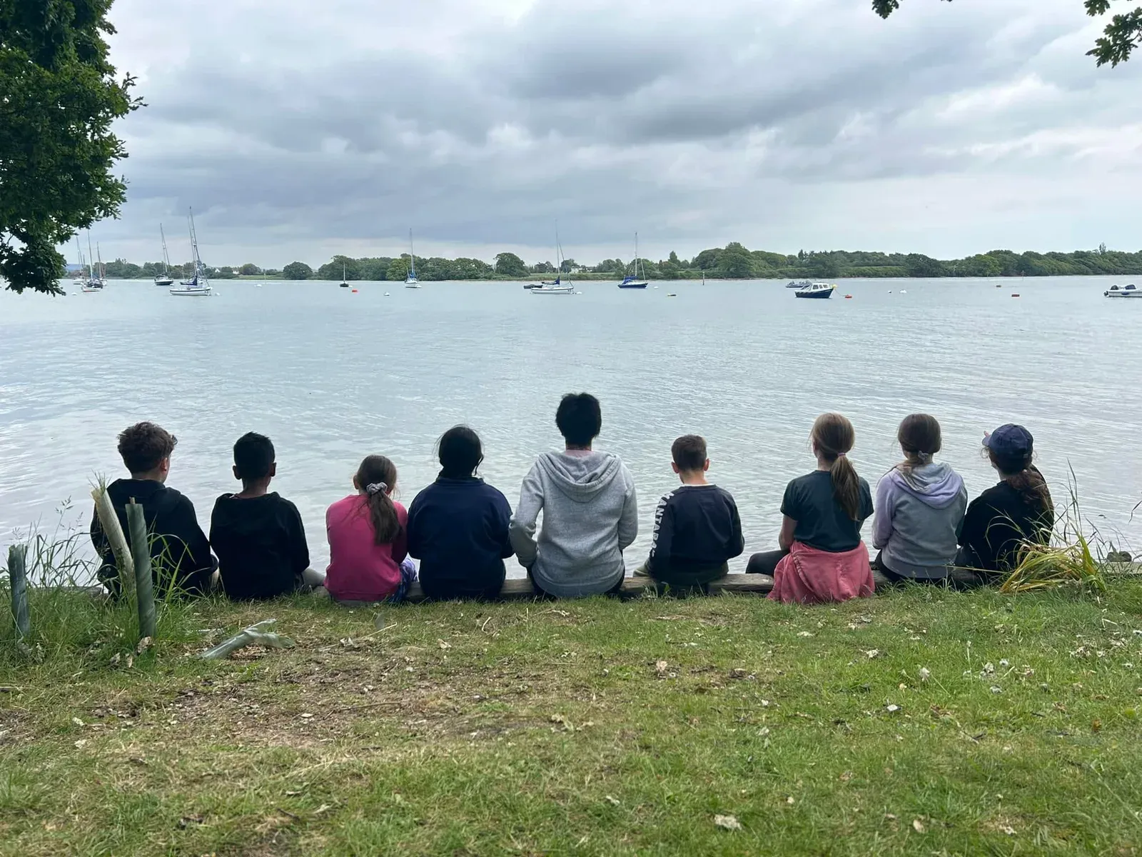 A class of pupils sitting in a row on the grass looking out over a lake on a school trip