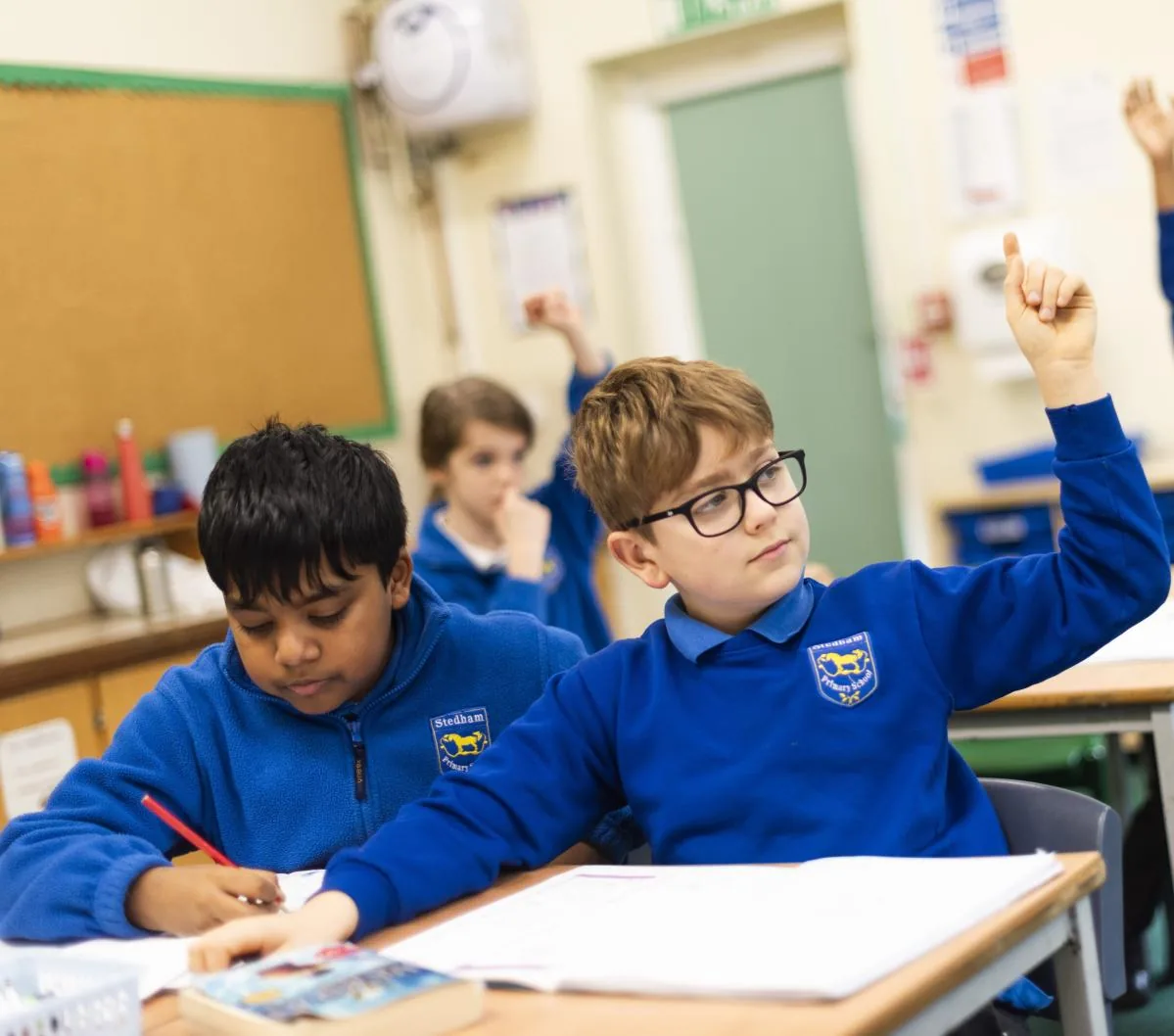 Two pupils in blue school jumpers with their hands up in class